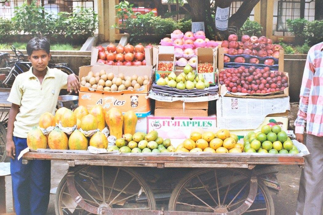 A boy standing beside a fruit cart loaded with mangoes and other fruit in Bombay