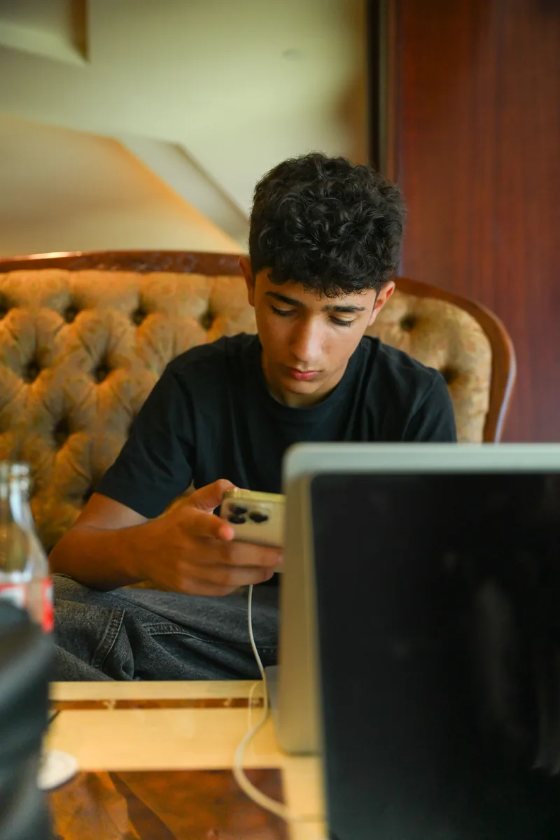 A teenage boy using a phone beside an open laptop
